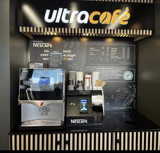 The same Ultracafé counter seen whole: the lit sign, the coffee machine, the price board — and our payment device mounted on the counter, where a customer meets it
